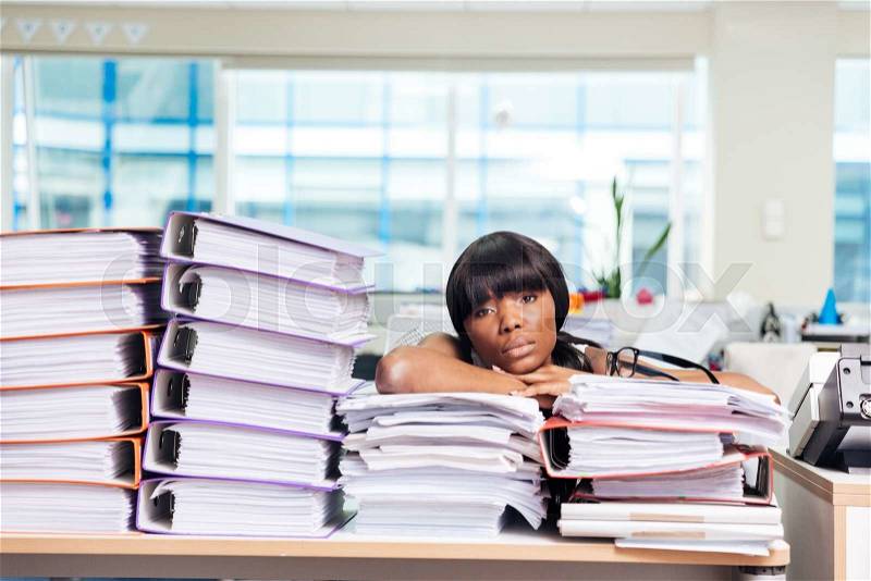 Tired afro american businesswoman sitting at the table with many work in office and looking at camera, stock photo
