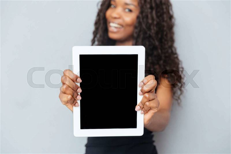 Smiling afro american woman showing blank tablet computer over gray background. Focus on tablet computer, stock photo