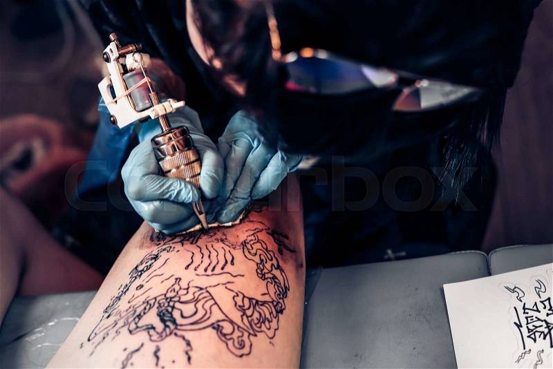 Closeup tattoo process in a professional salon. Master makes tattoo pattern on the client leg. Master works in sterile blue gloves, stock photo