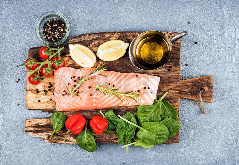 Ingredients for cooking healthy dinner. Raw salmon fillet, spinach, tomatoes, olive oil, lemon, peppers, rosemary and spices on a rustic wooden board over concrete textured grey background. Top view, stock photo