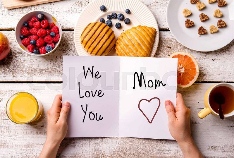 Mothers day composition. Hands of unrecognizable woman holding greeting card with We love you, Mom, text. Breakfast meal. Studio shot on wooden background, stock photo