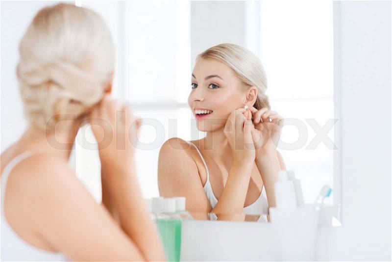 Beauty, jewelry and people concept - smiling young woman trying on diamond earring and looking to mirror at home bathroom, stock photo
