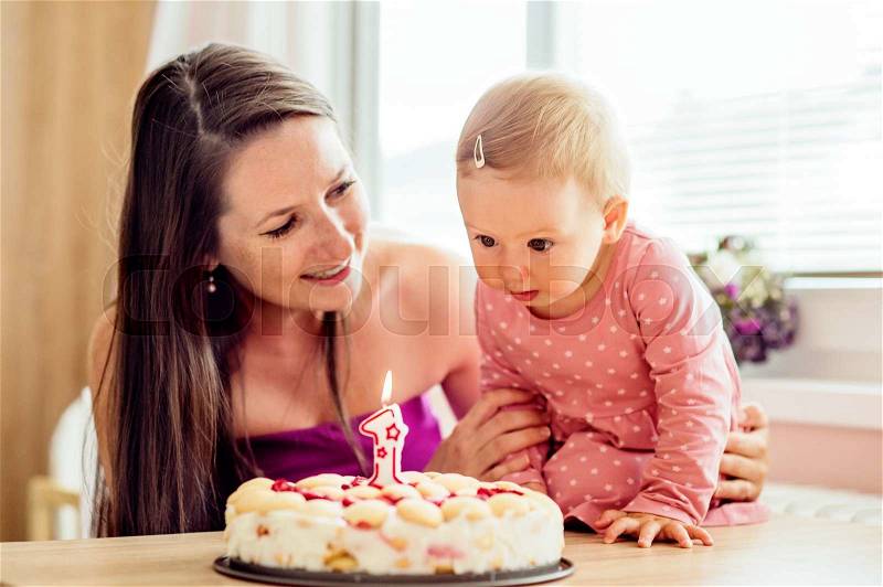 Young mother holding her cute baby daughter with birthday cake, blowing candle, stock photo