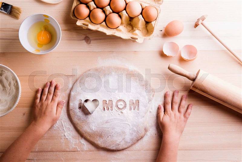 Mothers day composition. Hands of unrecognizable girl baking cookies. I love Mom sign made of flour and cookie cutter. Wooden background, stock photo
