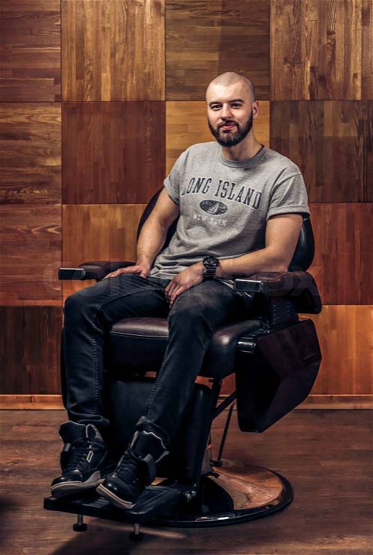 Happy client after beard haircut. Enjoying his perfect beard. Handsome young bearded man looking at the camera and sitting in a chair on a wooden texture, stock photo