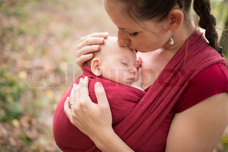 Mother carrying her cute baby daughter in sling, kissing her, outside in autumn nature, stock photo