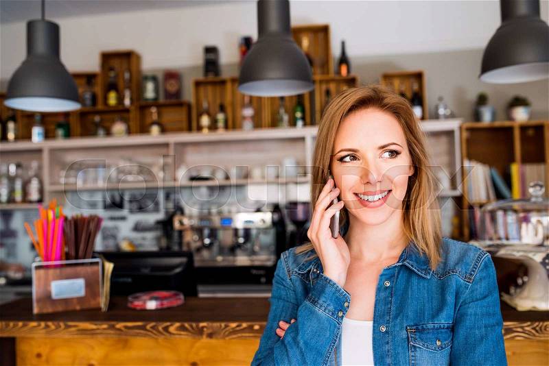 Blond woman in denim shirt standing at the bar in modern city cafe, making a phone call, stock photo