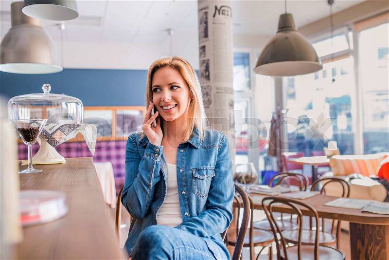 Blond woman in denim shirt sitting at the bar in modern city cafe, making a phone call, stock photo
