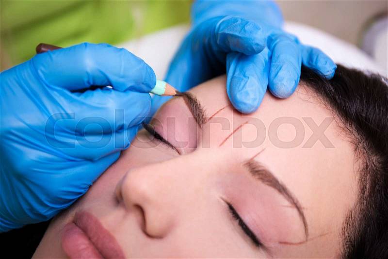 Close up of cosmetician preparing young woman for permanent eyebrow make up procedure, stock photo