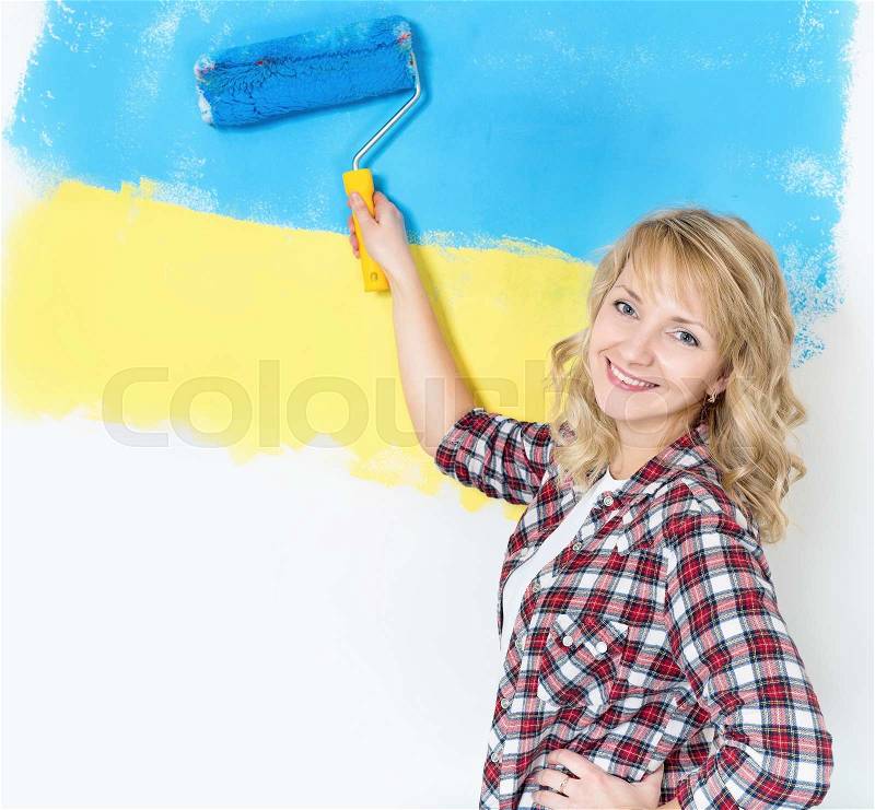 Happy woman makes repairs at home - painting wall at room. Portrait of smiling woman painting big Ukrainian flag on wall at home, stock photo