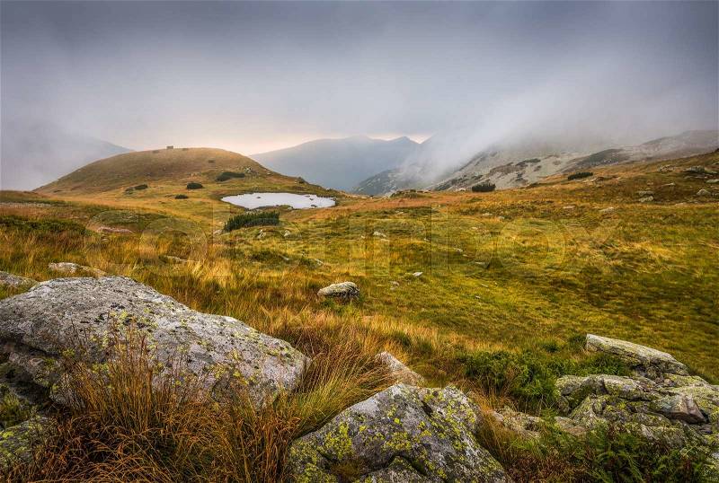 Foggy Mountain Landscape with a Tarn and Rocks in Foreground at Sunset, stock photo