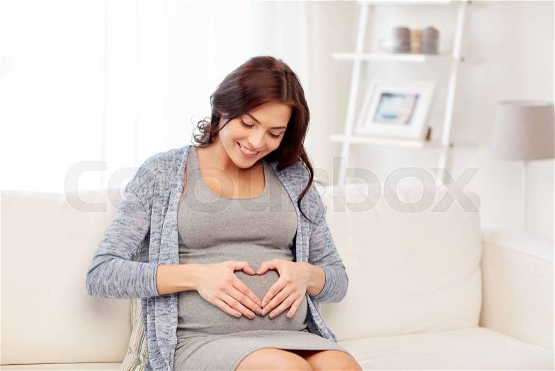 Pregnancy, love, people and expectation concept - happy pregnant woman sitting on sofa and making heart gesture at home, stock photo