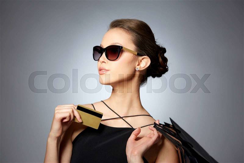 Sale, finances, fashion, people and luxury concept - happy beautiful young woman in black sunglasses with credit card and shopping bags over gray background, stock photo