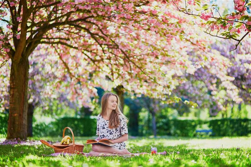 Girl having picnic and reading book in a beautiful cherry garden. Student preparing for exams or doing homework outdoors, stock photo