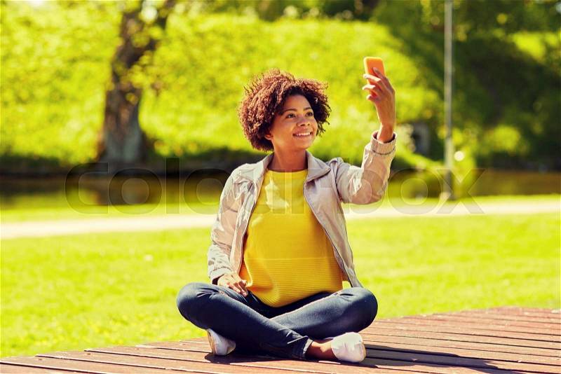 Technology, communication and people concept - happy african woman taking selfie with smartphone and sitting on bench in summer park, stock photo