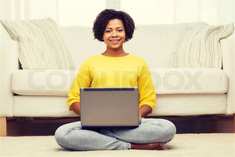 People, technology and leisure concept - happy african american young woman sitting on floor with laptop computer at home, stock photo