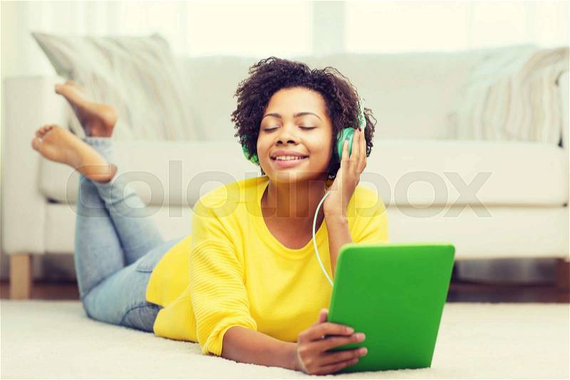People, technology and leisure concept - happy african american young woman lying on floor with tablet pc computer and headphones listening to music at home, stock photo