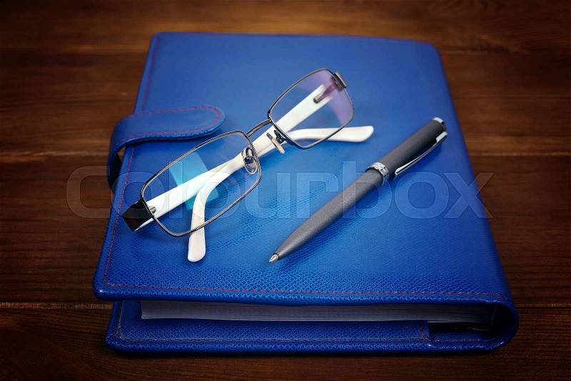 Blank blue leather diary, glasses and pen on wooden desk, stock photo