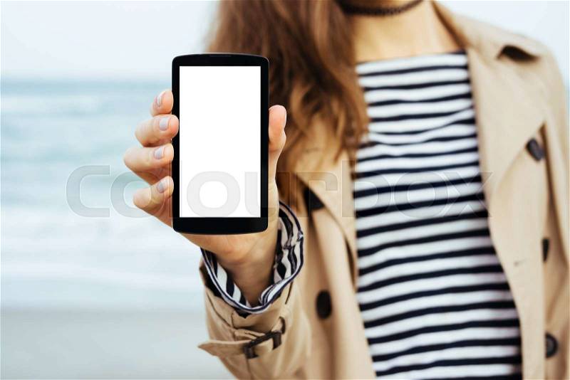 Girl in beige coat and striped t-shirt shows a blank screen phone on sea background, stock photo