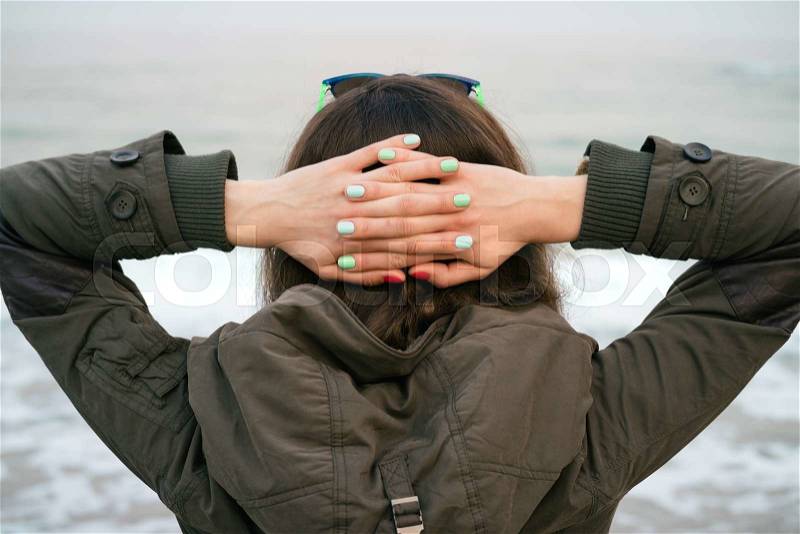 Girl in the green jacket with a hood looks on the sea put her hands on the back of the head, stock photo