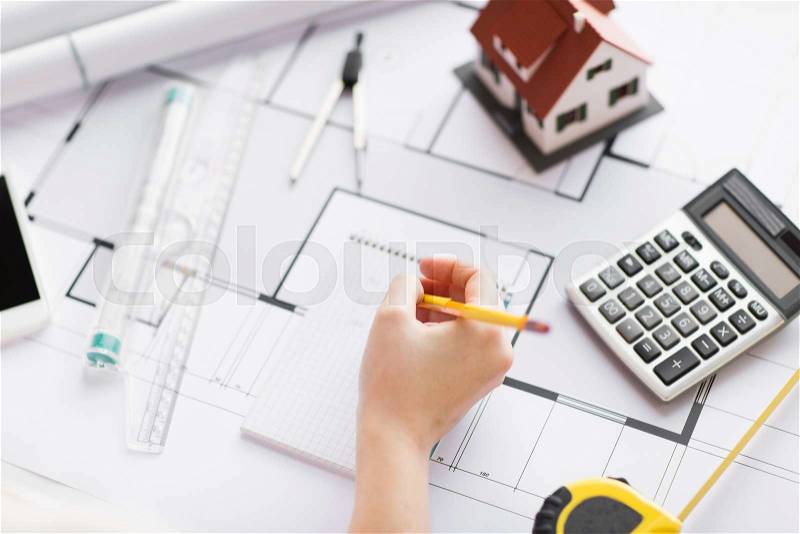 Business, architecture, building, construction and people concept - close up of architect hand with blueprint and architectural tools writing to notebook, stock photo