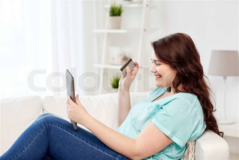 People, internet bank, online shopping, technology and e-money concept - happy young plus size woman sitting on sofa with tablet pc computer and credit card at home, stock photo