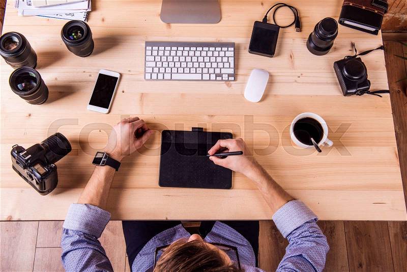 Photographer at the desk, wearing smart watch, working on graphic tablet camera. Computer, smart phone and various object lens around the workplace, stock photo