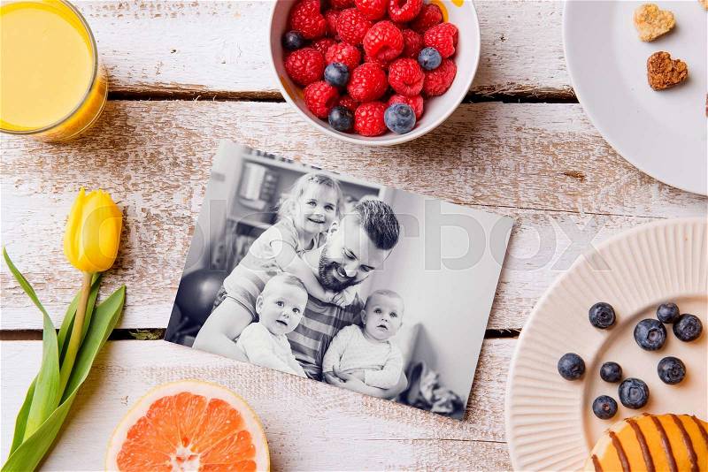 Fathers day composition. Black-and-white pictures of father holding her little daughters and a breakfast meal. Studio shot on white wooden background, stock photo