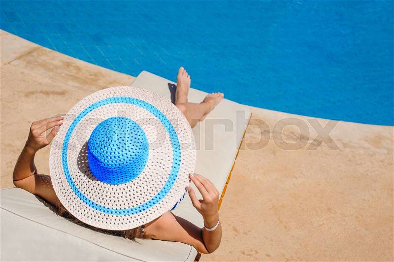 Happy woman with hat sunbathing on a sun lounger by the pool, stock photo