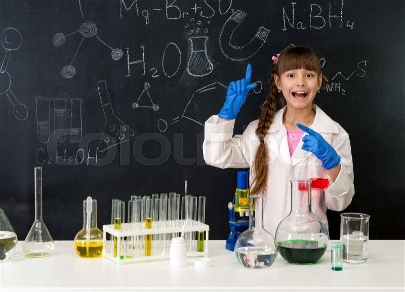Schoolgirl in chemistry lab in white gown pointing at formula on blackboard near table with reagents, stock photo