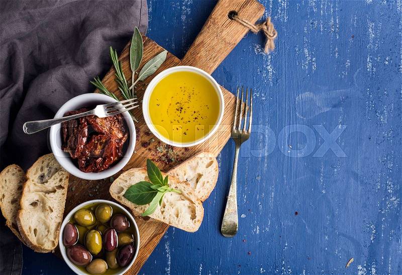 Mediterranean snacks set. Olives, oil, herbs and sliced ciabatta bread on yellow rustic oak board over painted dark blue background, top view, copy space, stock photo