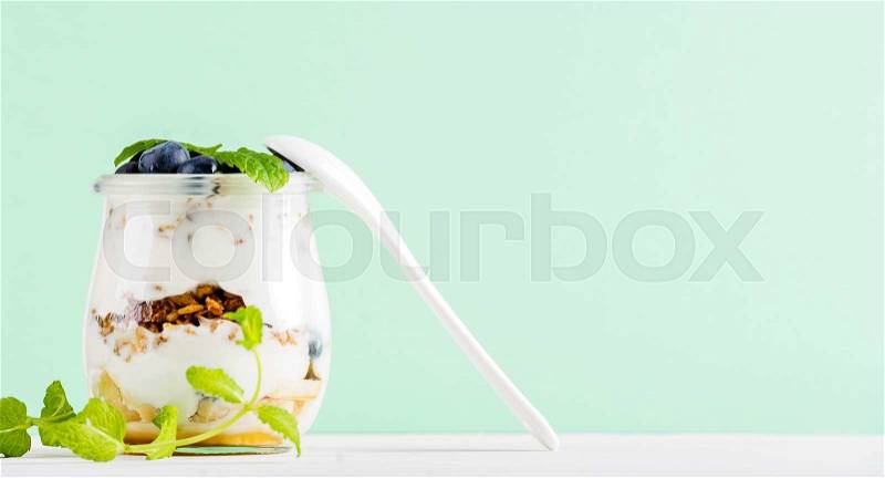 Yogurt oat granola with jam, blueberries and green leaves in glass jar on pastel mint backdrop, copy space, stock photo