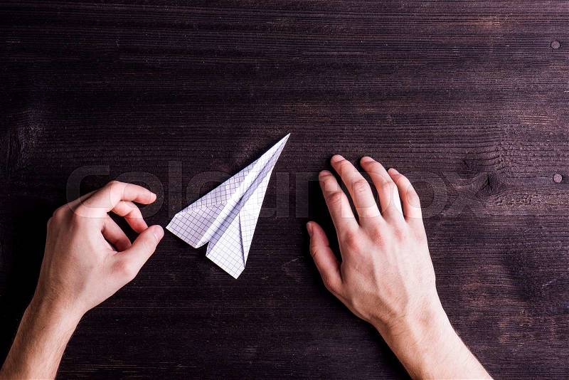 Office desk with hands of unrecognizable man holding paper airplane. Flat lay. Workplace. Studio shot on dark wooden background, stock photo
