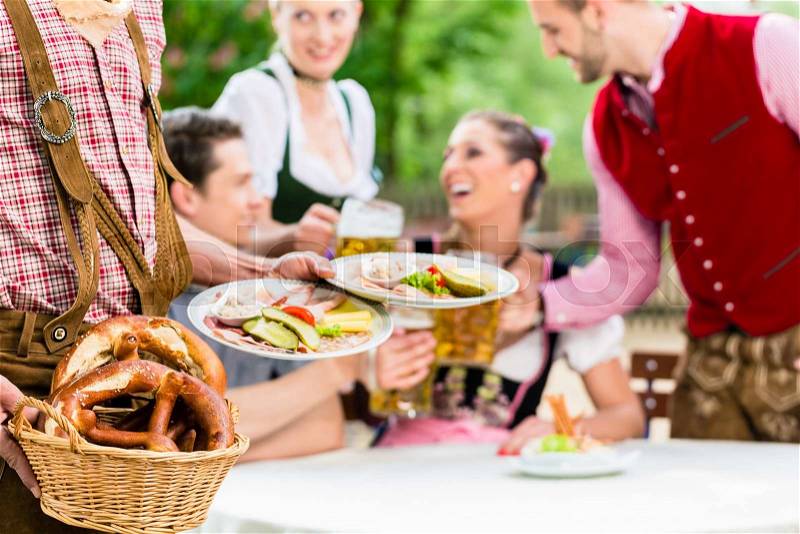 Waiter serving food in Bavarian beer garden, people eating and drinking in background, stock photo