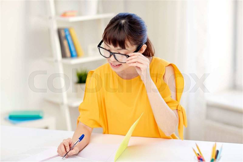 People, education, high school and learning concept - happy asian young woman student in glasses reading book and writing to notepad at home, stock photo