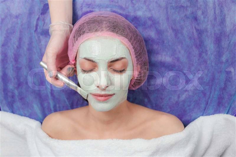 Close-up portrait of beautiful girl applying facial clay mask and beauty treatments lying on a table in spa beauty salon. Cosmetician applying facial beauty mask for young beautiful woman at spa salon, stock photo