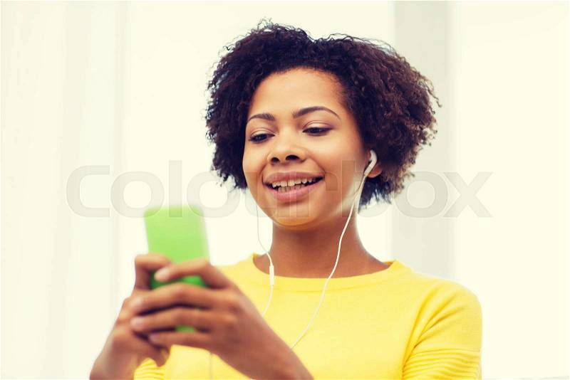 People, technology and leisure concept - happy african american young woman sitting on sofa with smartphone and earphones listening to music at home, stock photo