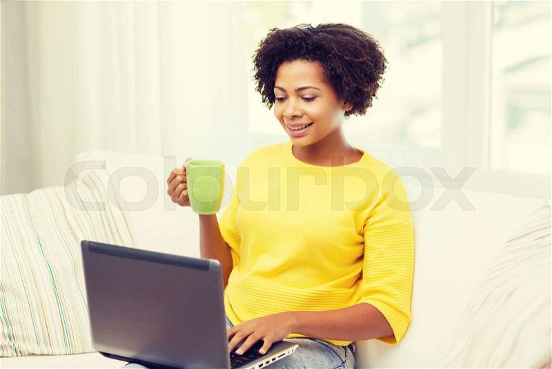 People, technology and leisure concept - happy african american young woman sitting on sofa with laptop computer and drinking tea from cup at home, stock photo