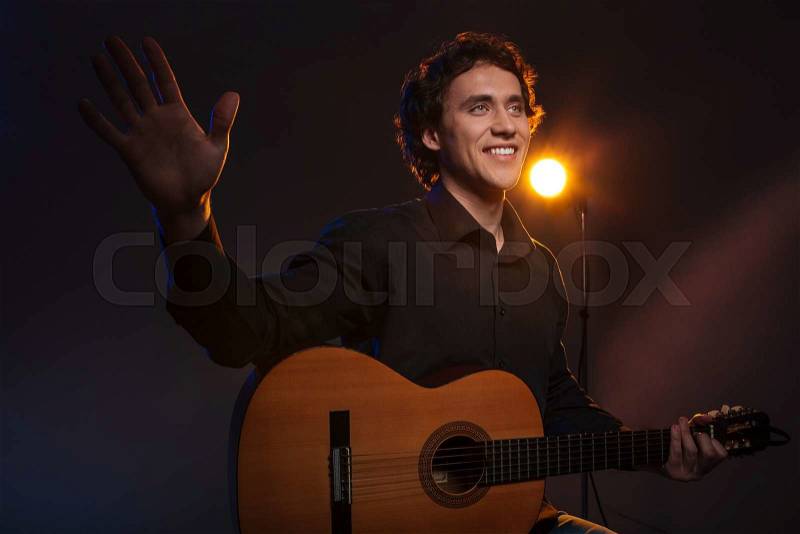 Happy man holding guitar and waving hand over dark background, stock photo