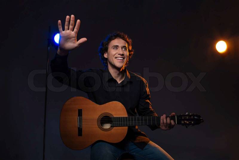 Smiling man holding guitar and waving hand over dark background, stock photo