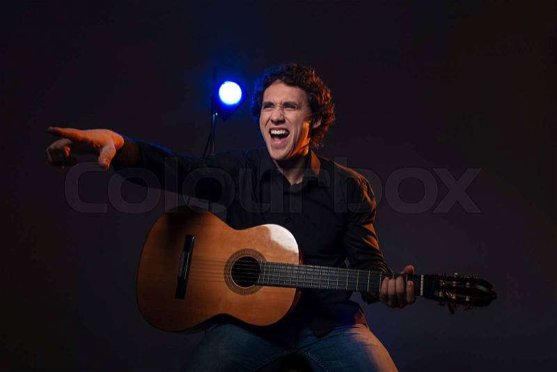 Happy man with guitar pointing finger on somebody over dark background, stock photo