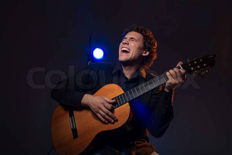 Cheerful man playing on guitar over dark background, stock photo