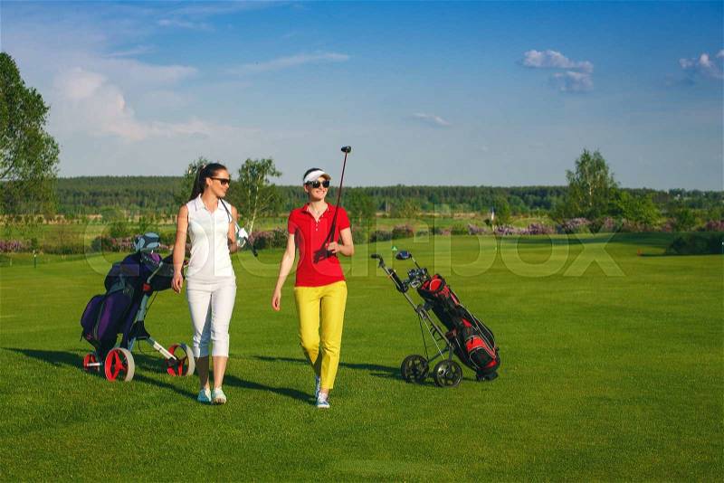 Two smiling sportive women golfers walking on golf course at sunny day, stock photo