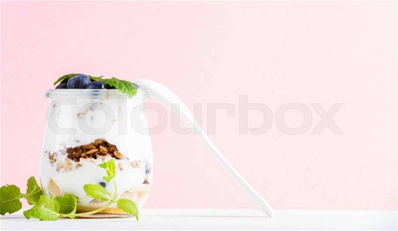 Yogurt oat granola with jam, blueberries and green leaves in glass jar on pastel pink backdrop, copy space, stock photo
