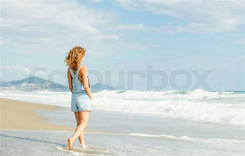 Young woman in blue denim jumpsuit walking along beach and the stormy ocean on sunny day, stock photo