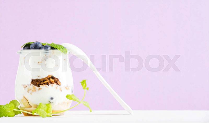 Yogurt oat granola with jam, blueberries and green leaves in glass jar on pastel lilac backdrop, copy space, stock photo