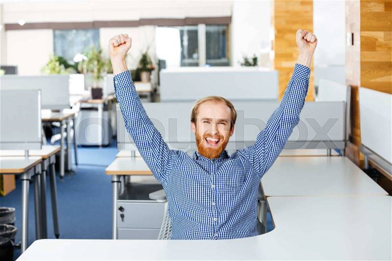 Happy excited bearded businessman sitting with raised hands and celebrating success in office, stock photo