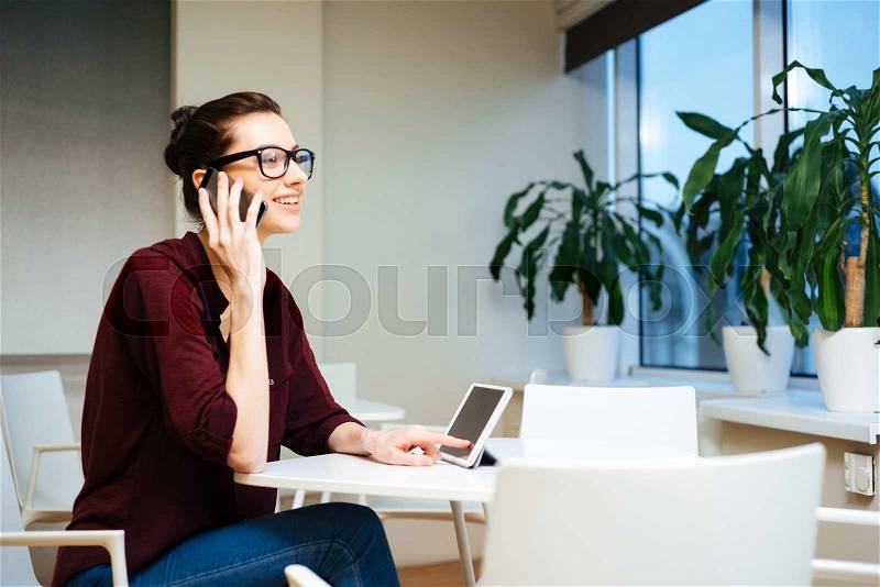 Cheerful attractive young busineswoman talking on cell phone and using tablet , stock photo