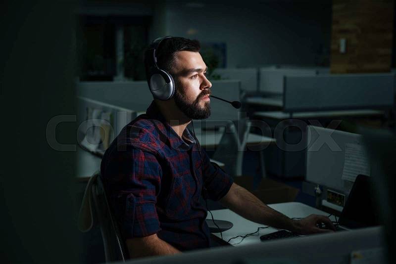 Concentrated handsome young man in headphones sitting and working in office in the evening, stock photo