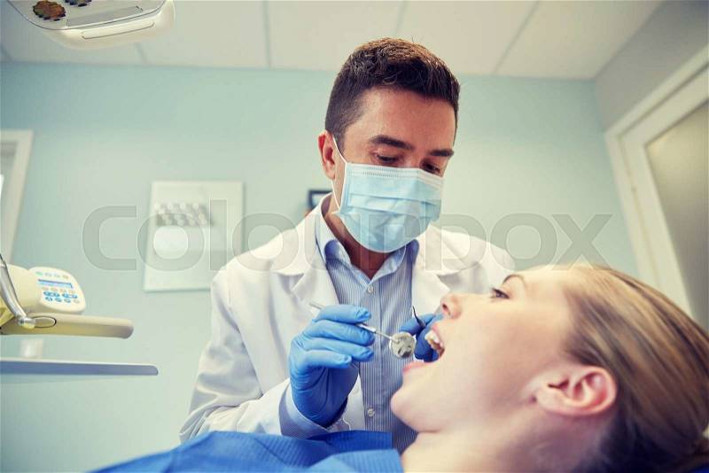 People, medicine, stomatology and health care concept - male dentist in mask with dental mirror and probe checking female patient teeth up at dental clinic office, stock photo